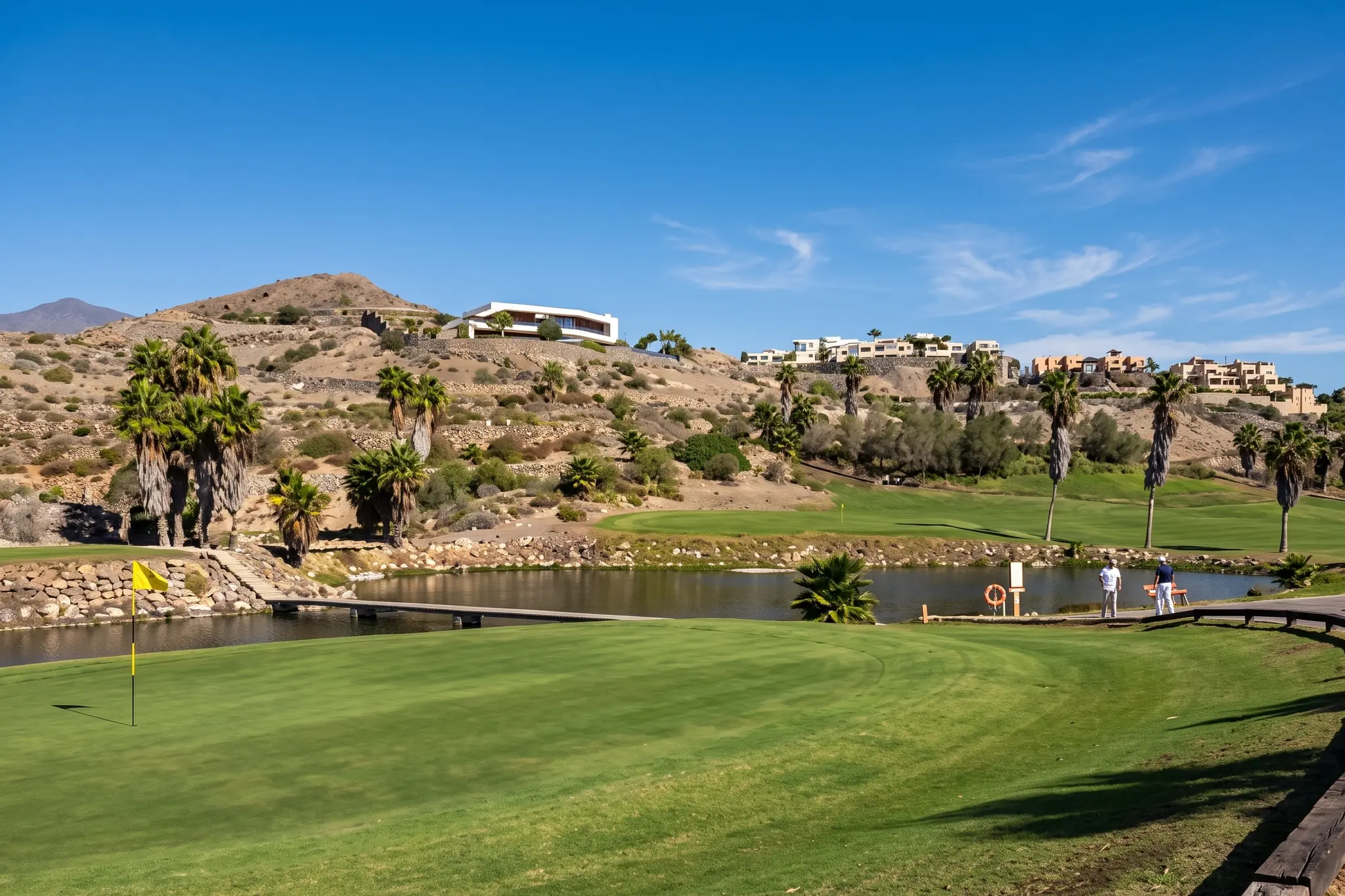 Green y lago de un campo de golf del sur de Gran Canaria, con palmeras y casas sobre la ladera al fondo.
