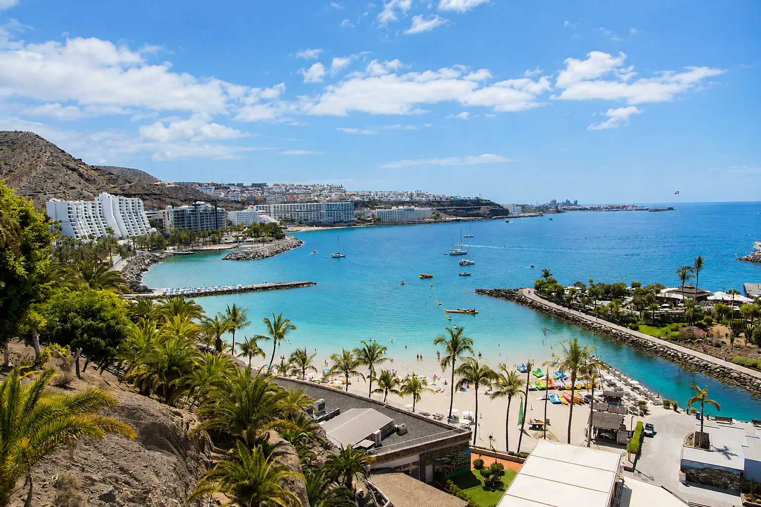 Bahía de arena clara y agua turquesa en la costa sur de Gran Canaria, con las palmeras del paseo y la montaña detrás.
