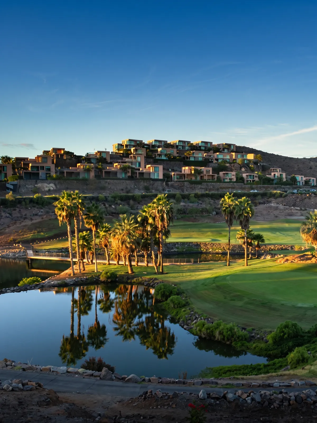 El campo de golf de El Salobre al atardecer, con sus casas escalonadas en la ladera, palmeras y un lago que las refleja.