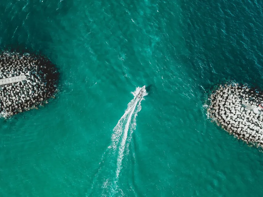 Una embarcación sale a mar abierto entre dos espigones, vista desde el aire sobre agua turquesa.