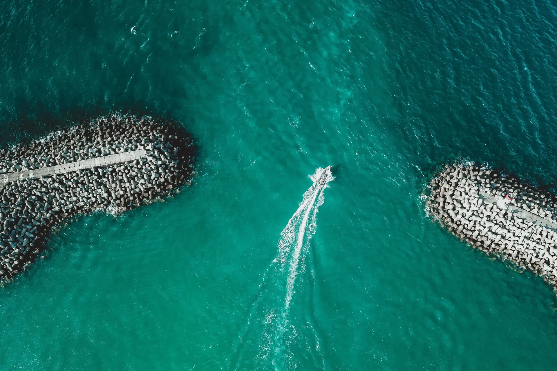 Una embarcación sale a mar abierto entre dos espigones, vista desde el aire sobre agua turquesa.
