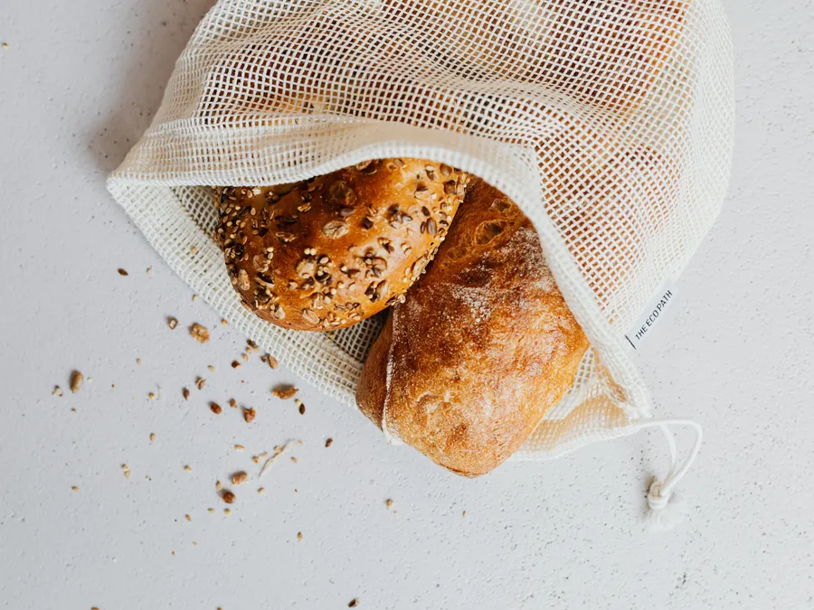 Dos panes recién horneados, uno de semillas y una chapata enharinada, dentro de una bolsa de malla de algodón.