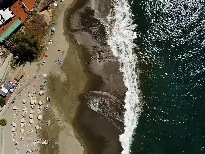Vista cenital de una playa: sombrillas y hamacas sobre la arena, la rompiente entrando en diagonal y el mar abierto a la derecha.
