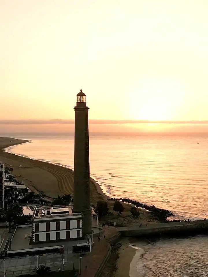 El faro de Maspalomas al amanecer, con la playa y el Atlántico en calma al fondo.