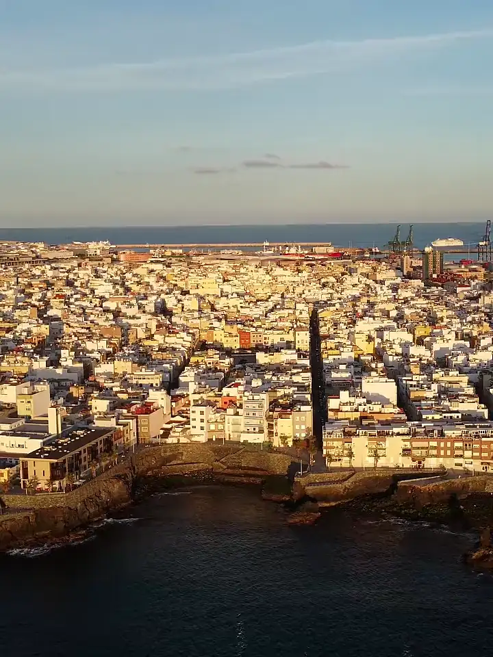 Vista aérea de Las Palmas de Gran Canaria al atardecer, con el caserío, el puerto y una cala rocosa en primer plano.