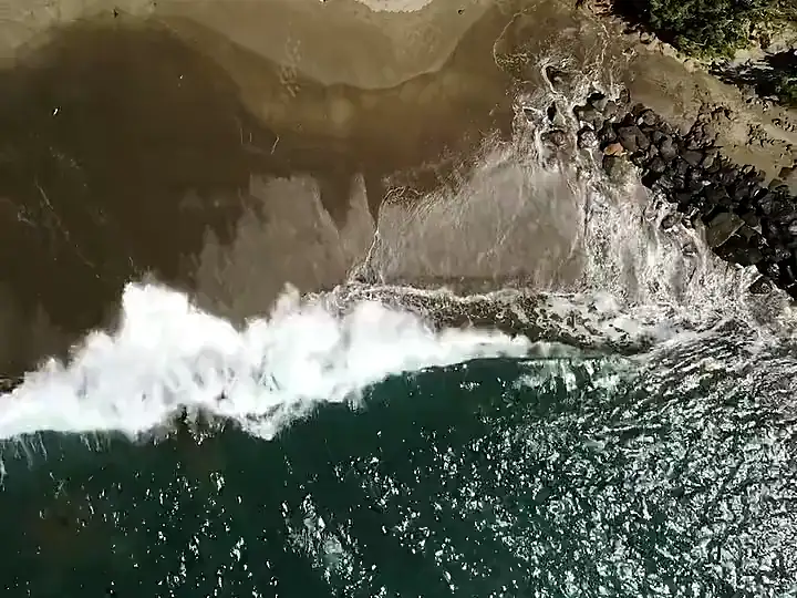 Vista cenital de la rompiente entrando sobre la arena y las rocas de una playa del sur de Gran Canaria.